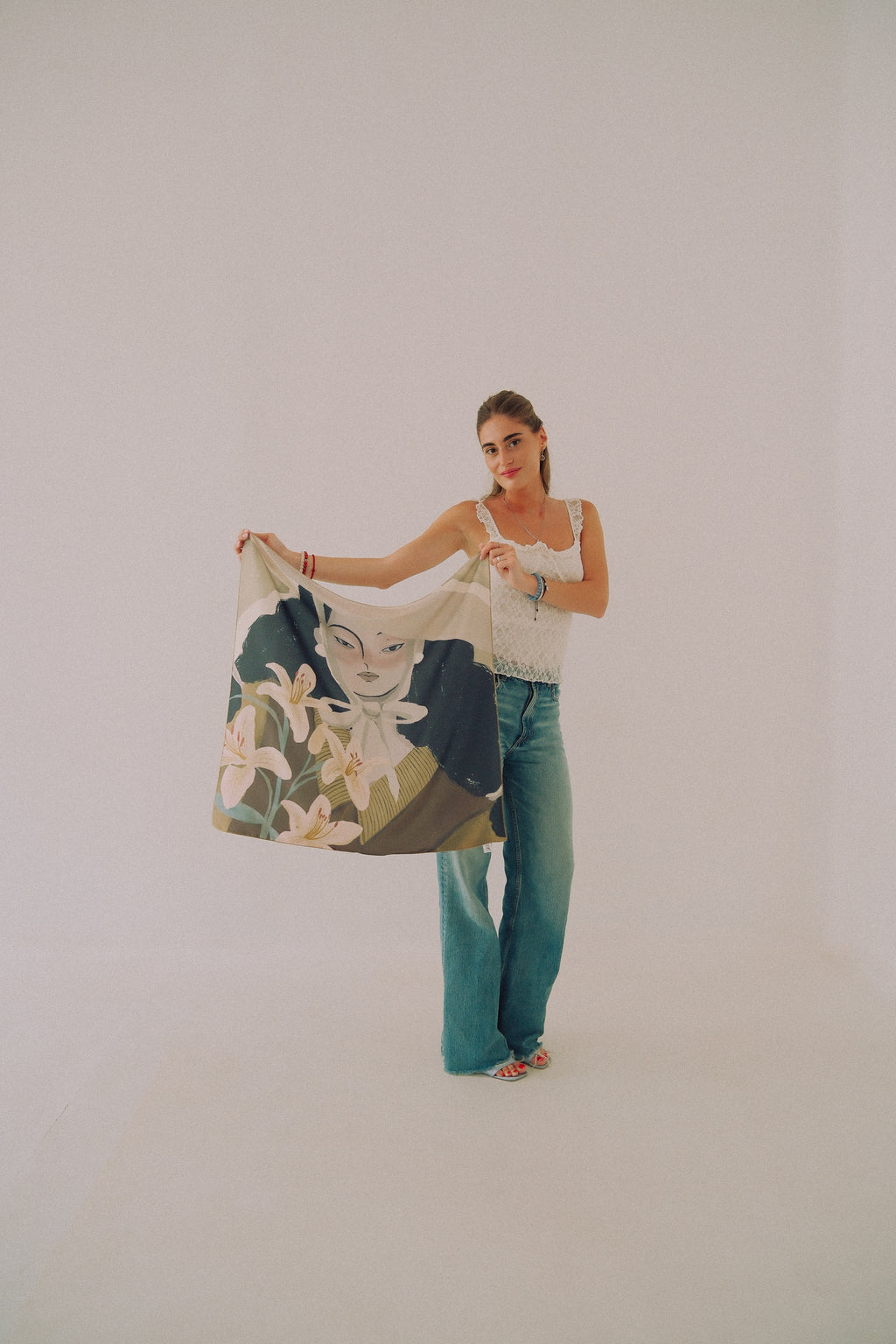A woman stands in a studio holding up a large square scarf or bandana featuring a vintage-style illustration of a woman's face and white flowers on a dark background. She is wearing a white sleeveless top and teal wide-leg jeans against a plain light gray backdrop.