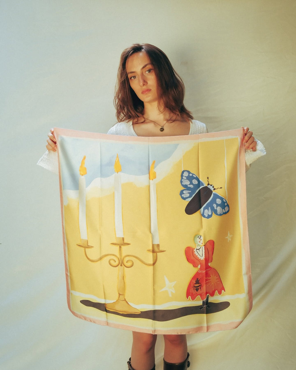 A woman holds up a square silk scarf featuring a whimsical illustration of a girl in a red dress, a blue butterfly, and a gold candelabra against a yellow background, with a pink border.