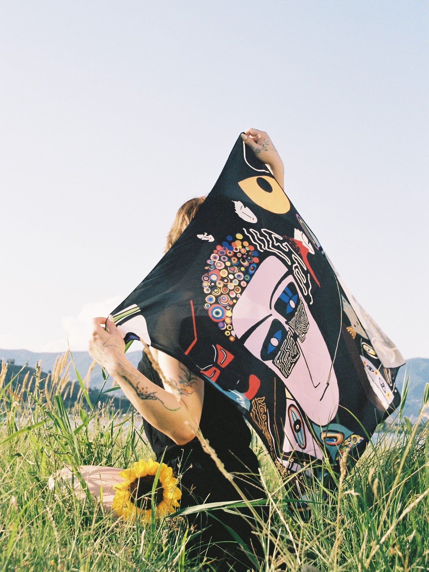 Person holding a colorful, abstract-patterned scarf in a grassy outdoor setting. Scarf by DAE x Nino Bakuradze