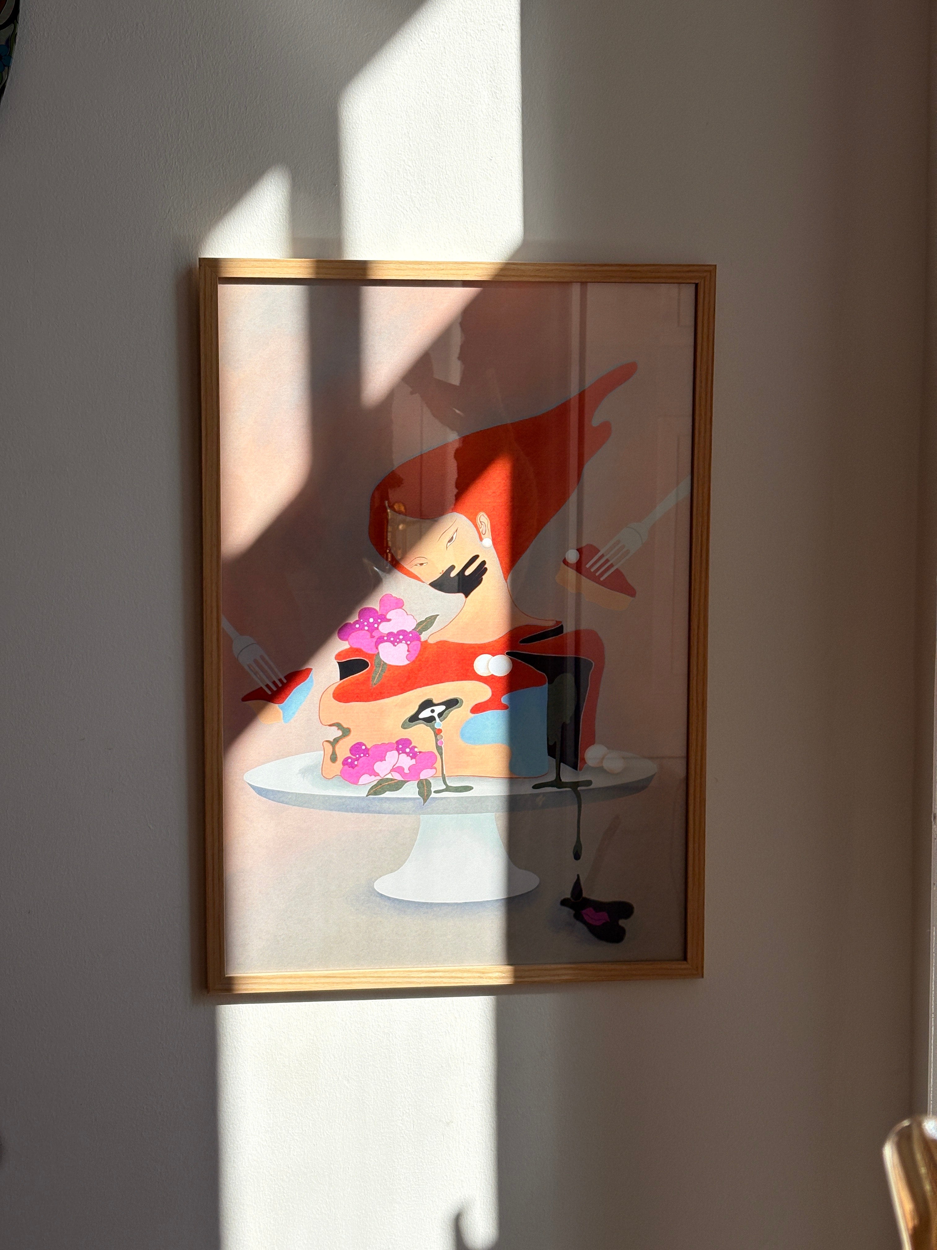 Framed artwork of a stylized character as a cake, flowers with her, on a white wall.