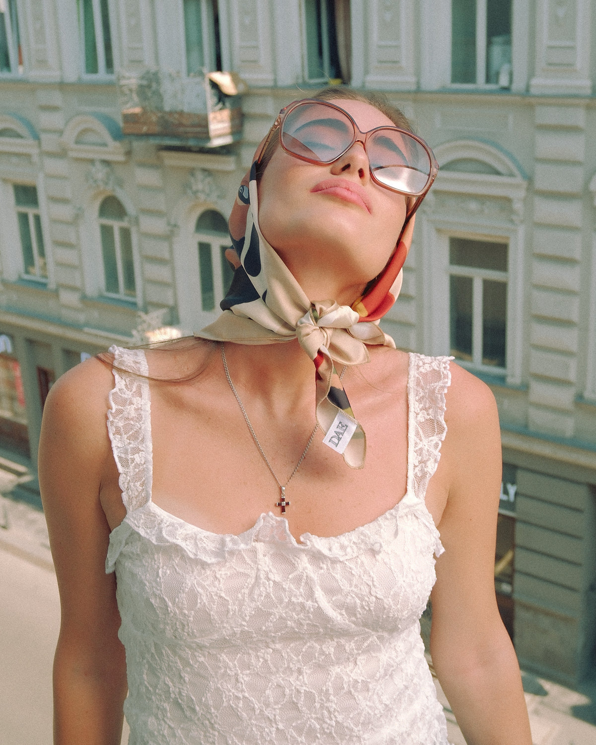 A woman wearing a white lace camisole top, a patterned silk neck scarf tied in a bow, oversized round sunglasses, and a delicate necklace, posing outdoors with a blurred historic building in the background. Wearing scarf by DAE