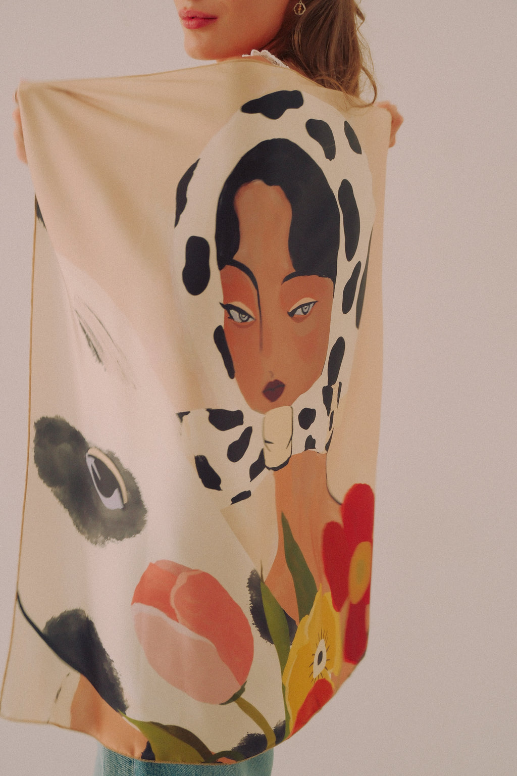 A woman holds up a large cream-colored silk scarf by DAE featuring a whimsical illustration of a woman in a black-and-white cow-print headscarf, with pink tulips and a red flower at the bottom.