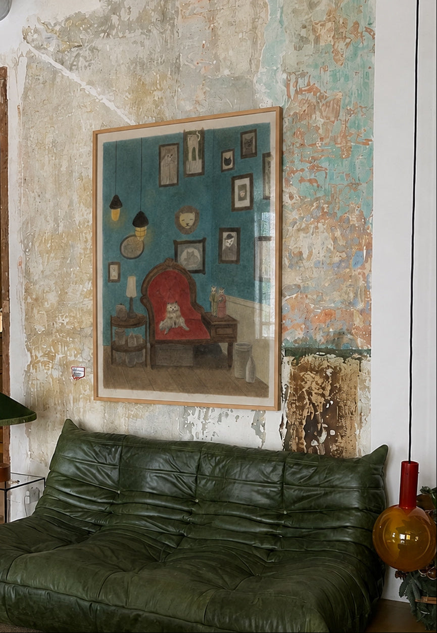 A framed art print depicting a cozy red armchair against a teal background, hanging on a heavily textured, peeling plaster wall above a dark green leather sofa in a rustic, industrial-style room.