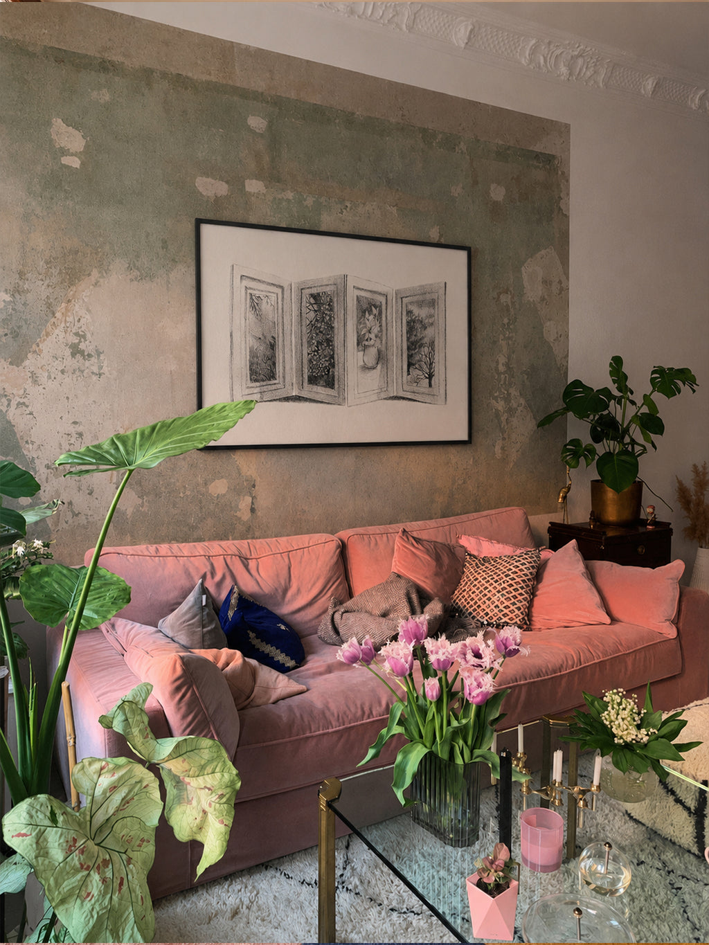 A dusty pink velvet sofa styled with pink and patterned throw pillows in a living room setting, set against a textured, distressed green-gray wall with a framed black and white print, poster by DAE hanging above it.