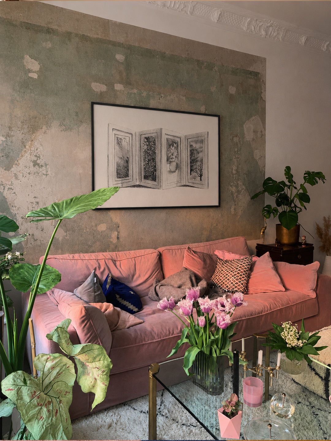 A dusty pink velvet sofa styled with pink and patterned throw pillows in a living room setting, set against a textured, distressed green-gray wall with a framed black and white print, poster by DAE hanging above it.