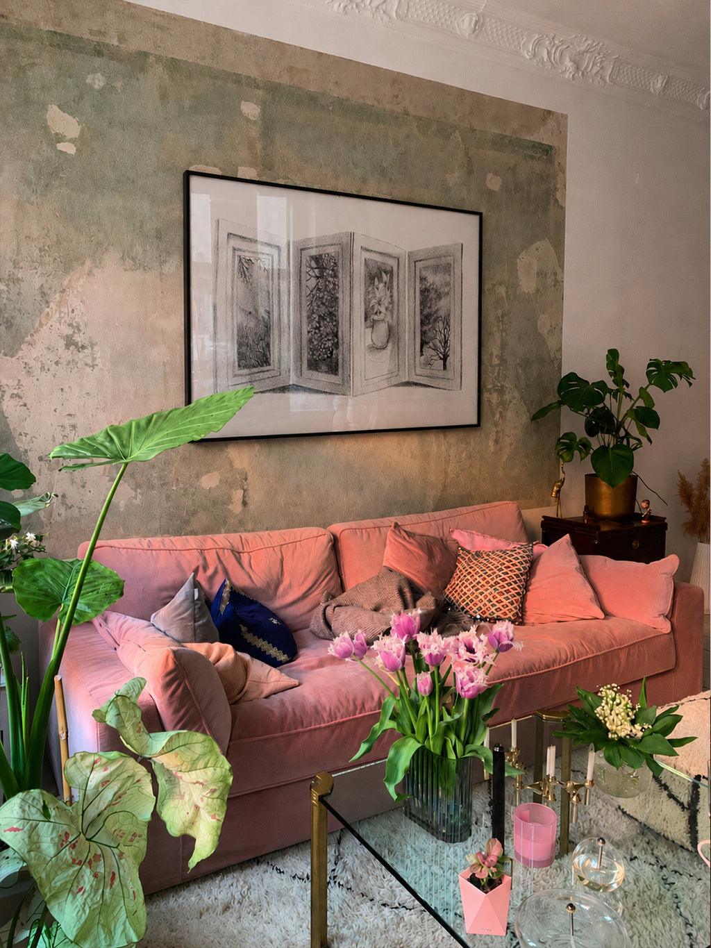 A pink velvet sofa in a styled living room setting, decorated with pink and neutral throw pillows, a glass coffee table with flowers, and a textured plaster wall with a framed black and white print.
