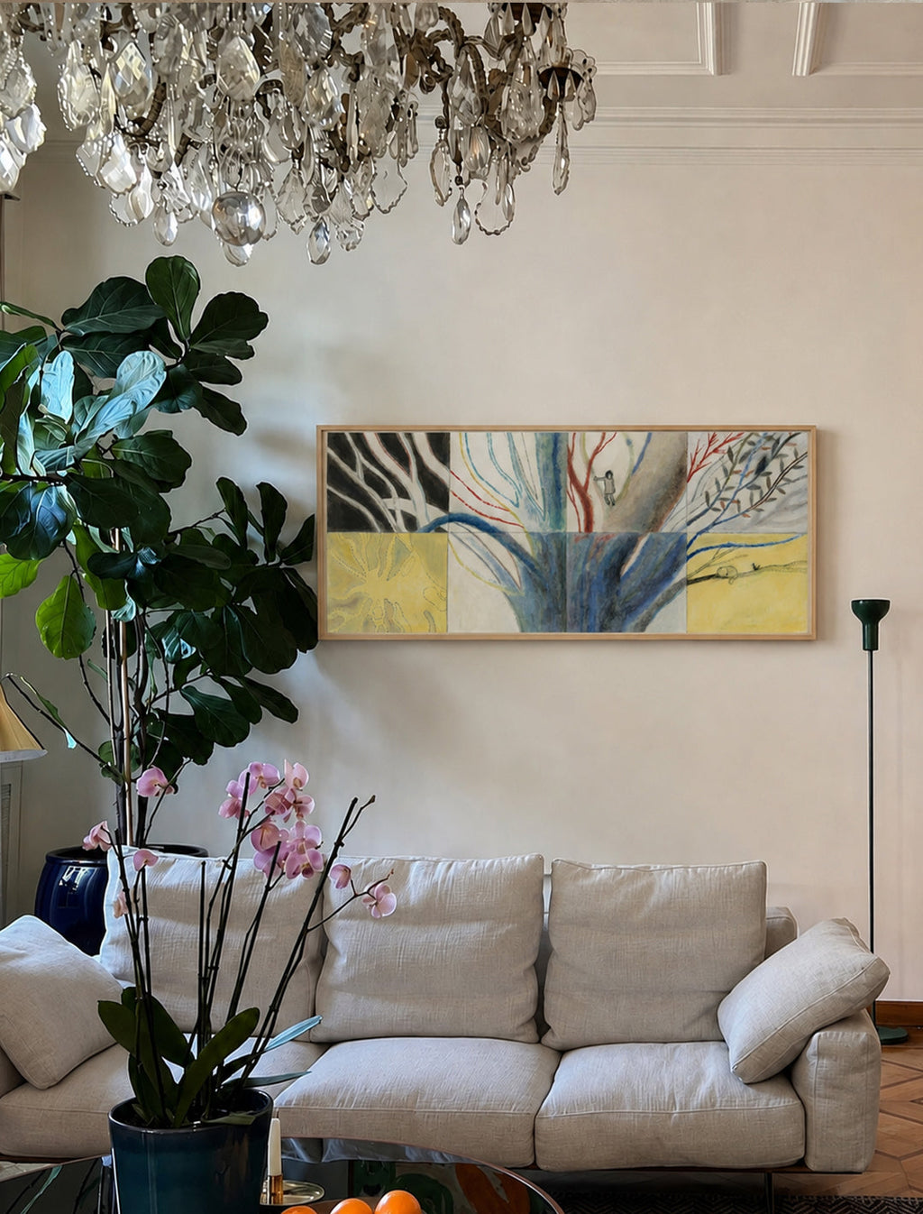 A framed abstract poster with blue, yellow, and black brushstrokes hangs on a cream wall above a light beige sofa in a styled living room with a chandelier, fiddle-leaf fig plant, and orchid.