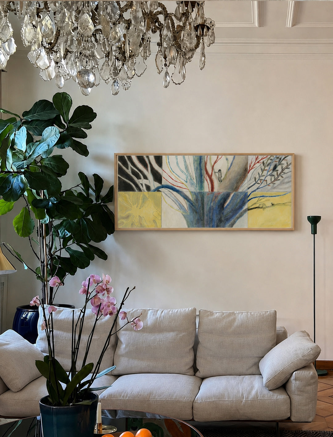 A framed abstract poster with blue, yellow, and black brushstrokes hangs on a cream wall above a light beige sofa in a styled living room with a chandelier, fiddle-leaf fig plant, and orchid.