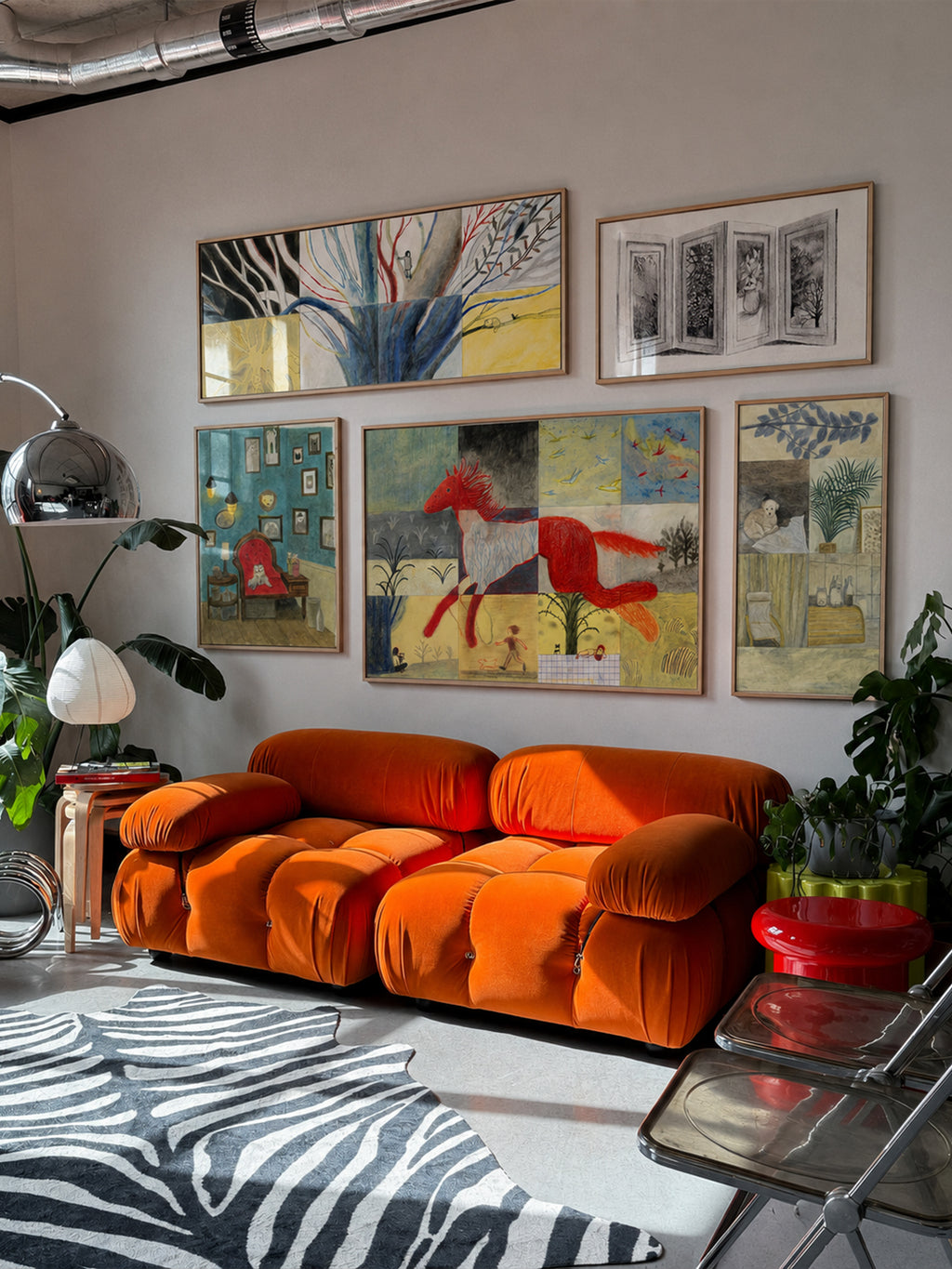 A modern living room featuring a prominent bright orange modular sofa, styled with a black and white zebra-patterned rug, a silver floor lamp, and a collection of colorful abstract paintings on a light gray wall.