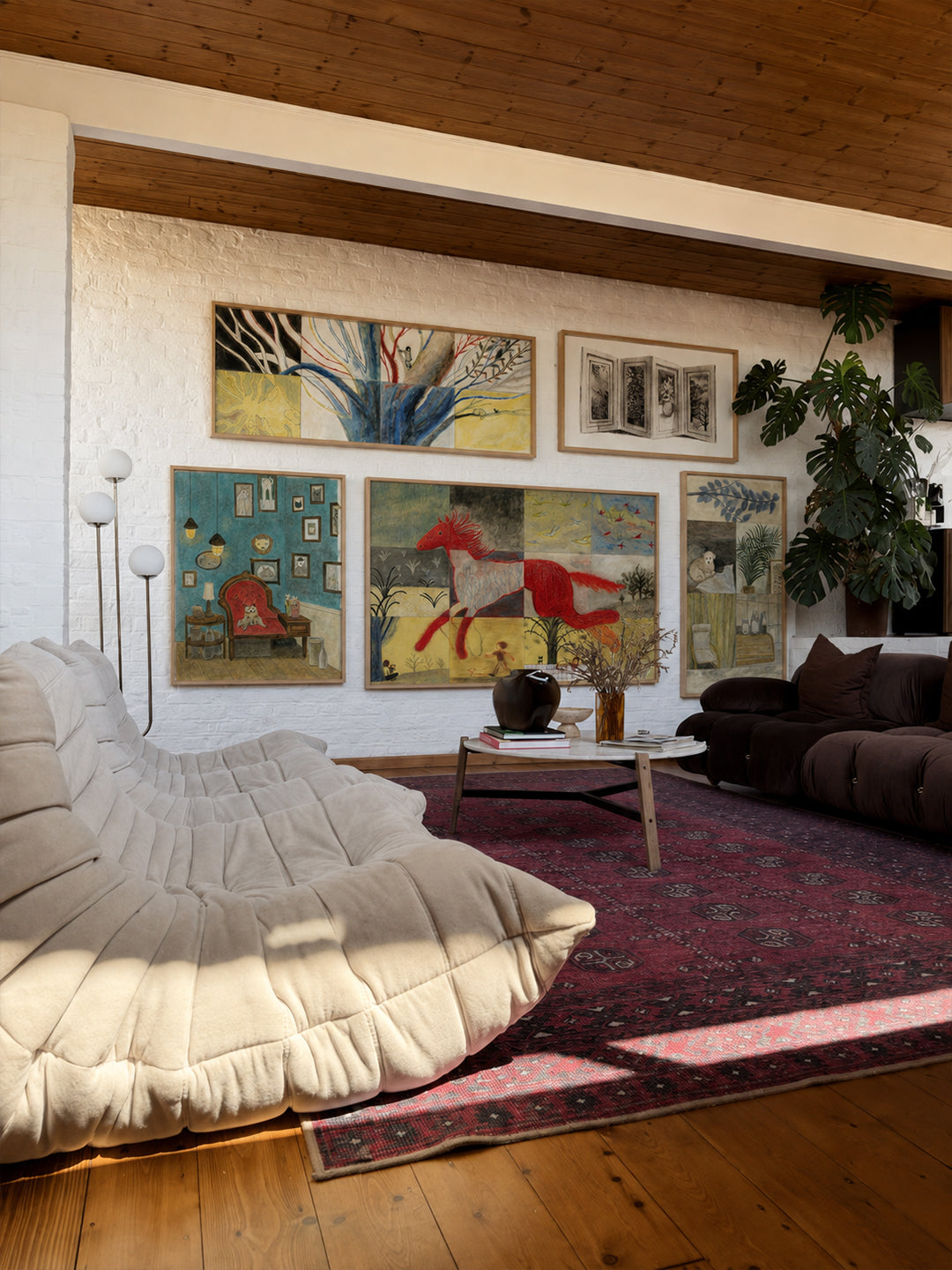 A styled living room interior featuring a large cream-colored modular daybed in the foreground, a dark brown sofa on the right, a red patterned rug, and a gallery wall of colorful abstract and figurative posters by DAE above.