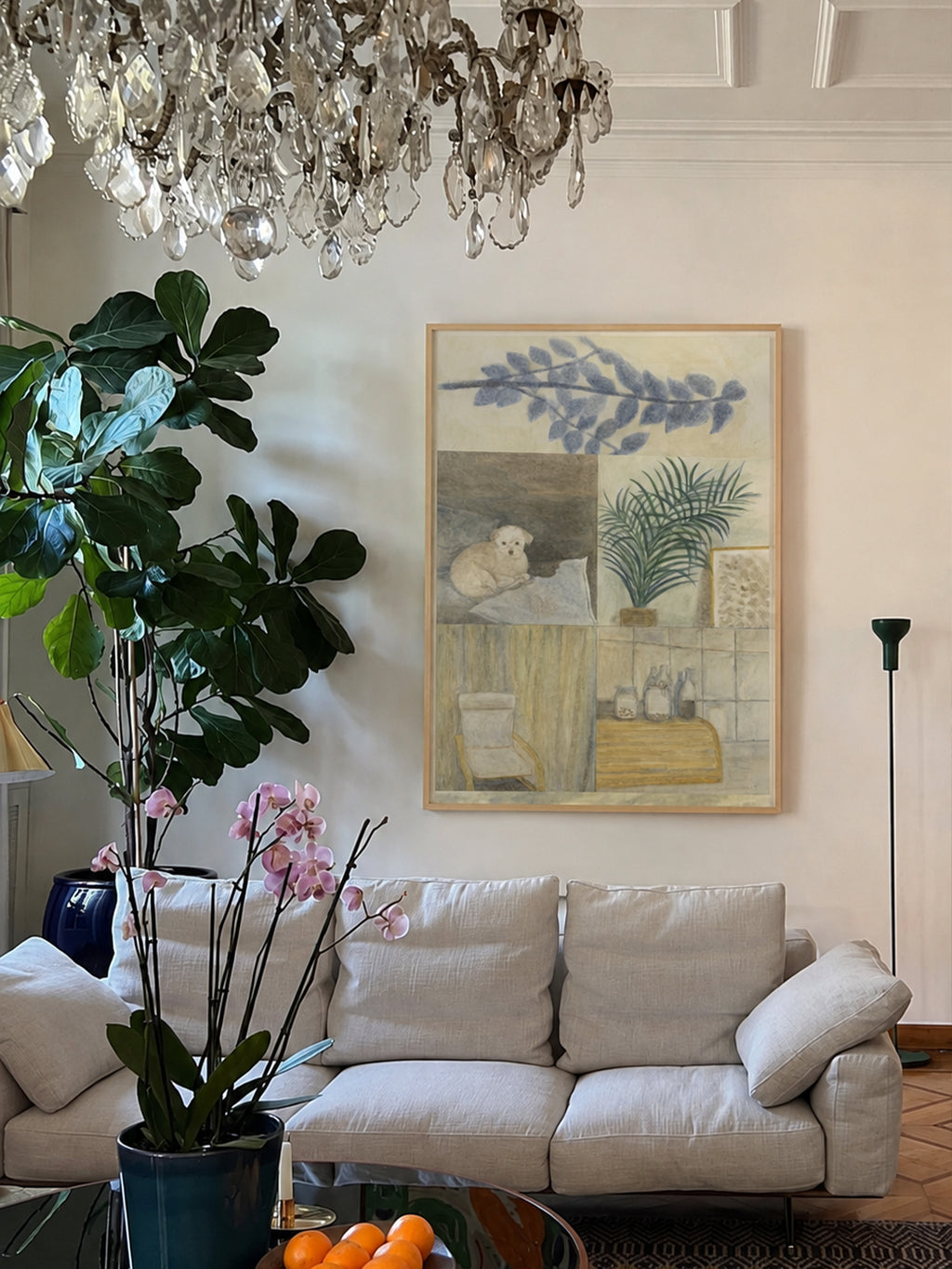 A framed botanical and interior art print by DAE hangs on a cream wall above a light beige sofa in a styled living room, with a large green plant, pink orchids, a crystal chandelier, and a floor lamp visible.