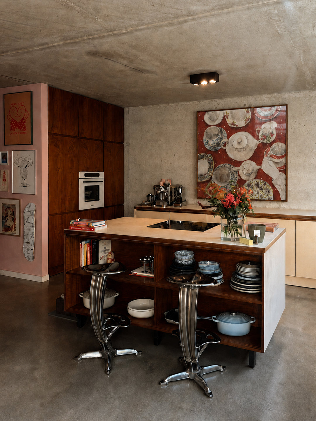 A modern kitchen island with a wood and white structure, featuring two chrome bar stools, open shelving with cookware, and a vase of flowers. The room has textured concrete floors and ceiling, a pink accent wall with framed art, poster by DAE and a large red and white abstract painting on the back wall.