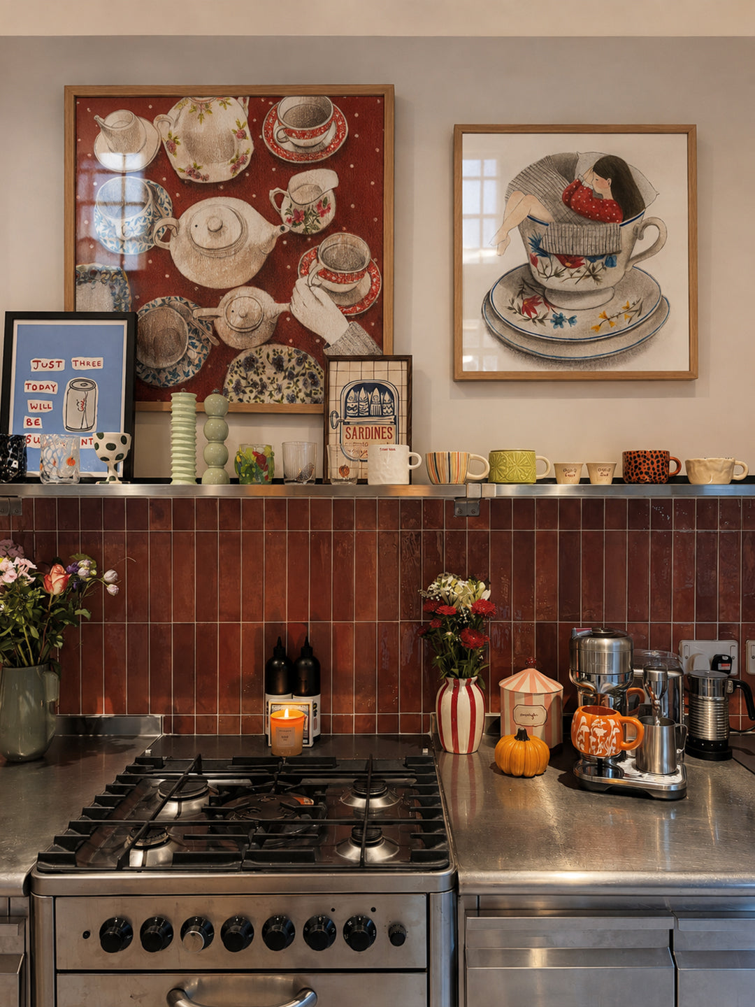 A styled kitchen interior with a stainless steel gas stove, dark wood subway tile backsplash, and two framed posters by DAE hanging on the wall above the counter.