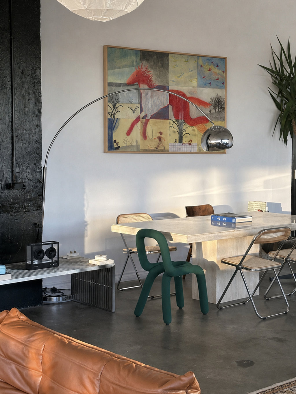 A modern dining room scene featuring a large abstract art print with red, blue, and yellow tones hanging on a white wall. The room includes a light wood dining table, a dark green molded chair, a silver floor lamp, and a brown leather sofa in the foreground.