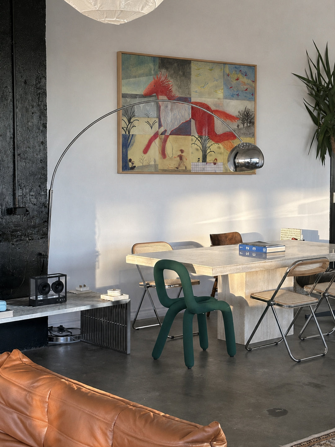 A modern dining room scene featuring a large abstract art print with red, blue, and yellow tones hanging on a white wall. The room includes a light wood dining table, a dark green molded chair, a silver floor lamp, and a brown leather sofa in the foreground.