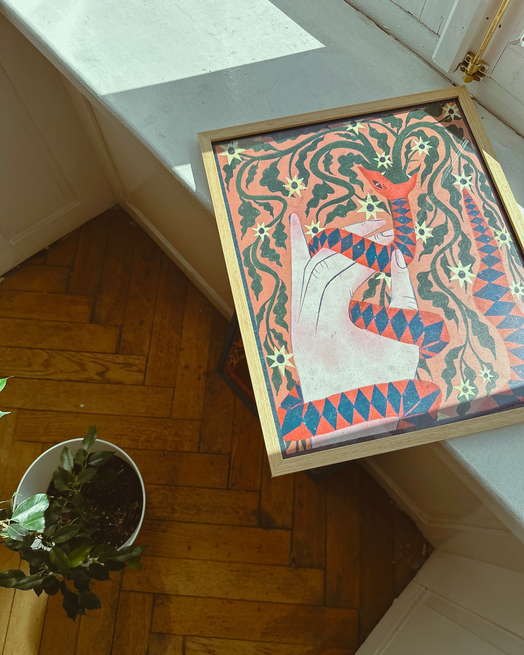 A poster with a geometric snake pattern in red, blue, and green colors displayed on a wooden floor next to a plant pot.