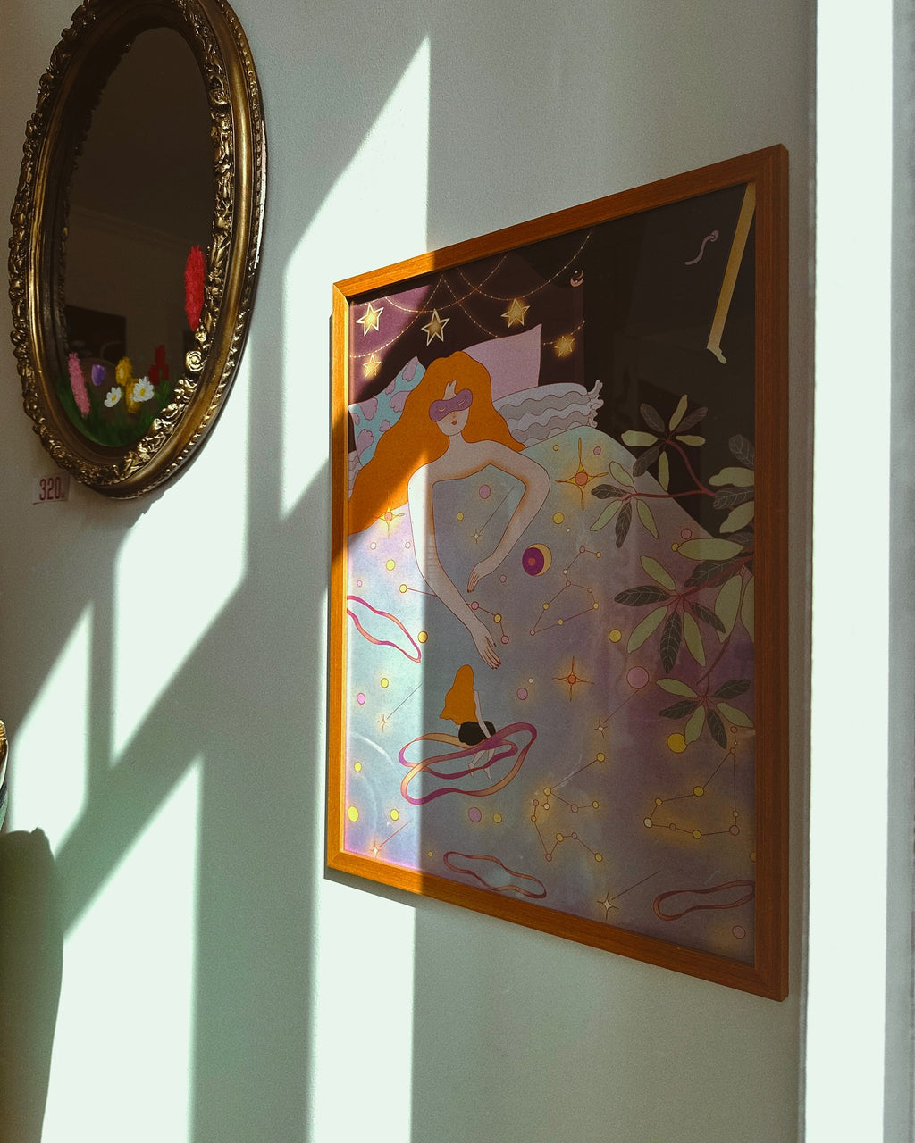 poster on the wall, with sun shining on it, next to mirror. illustration by leah han which represents woman in bed, laying with stars