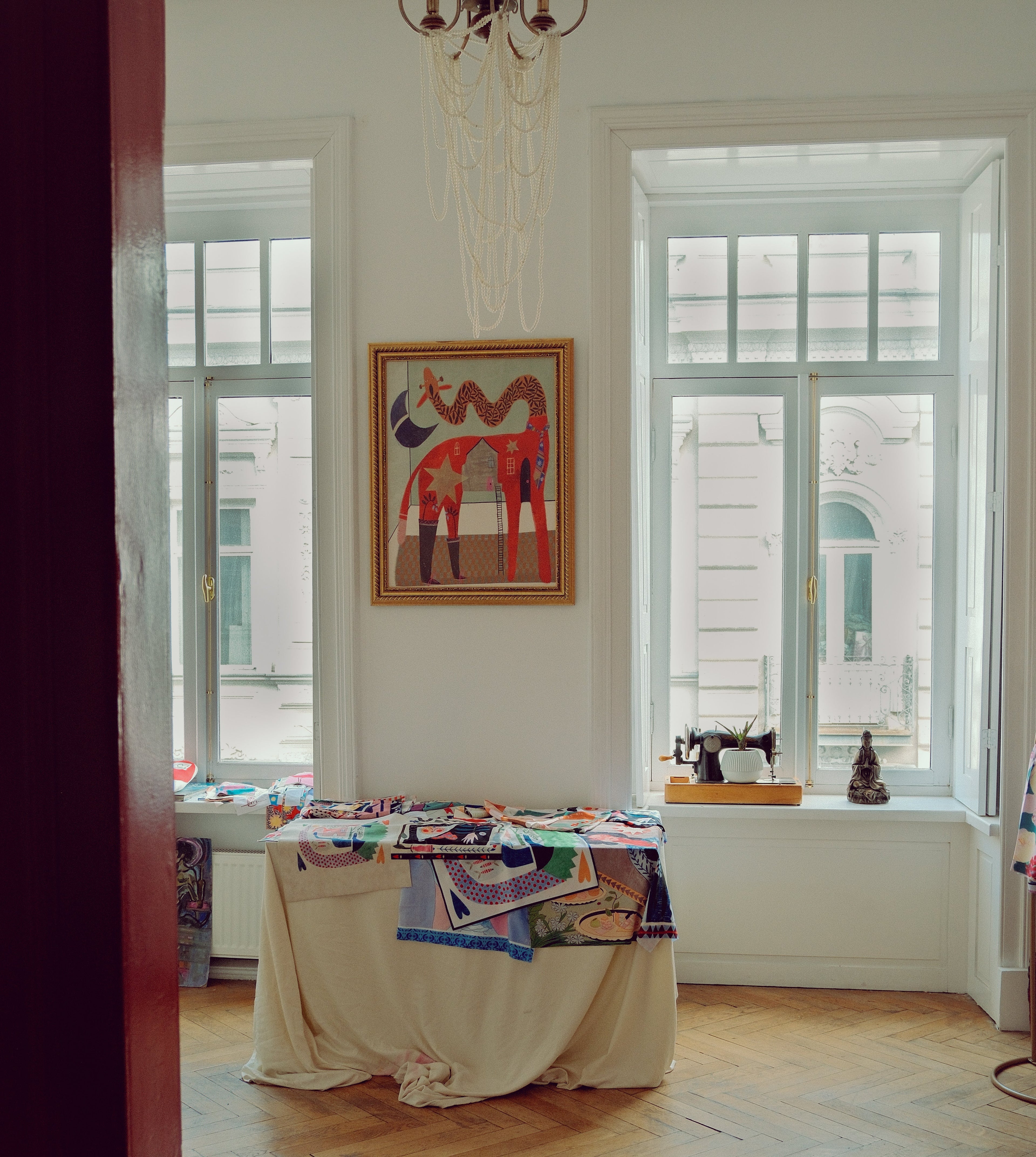 A framed colorful painting of stylized horses hangs on a white wall in a bright room with large windows, a draped table covered in textiles, and a wood herringbone floor.