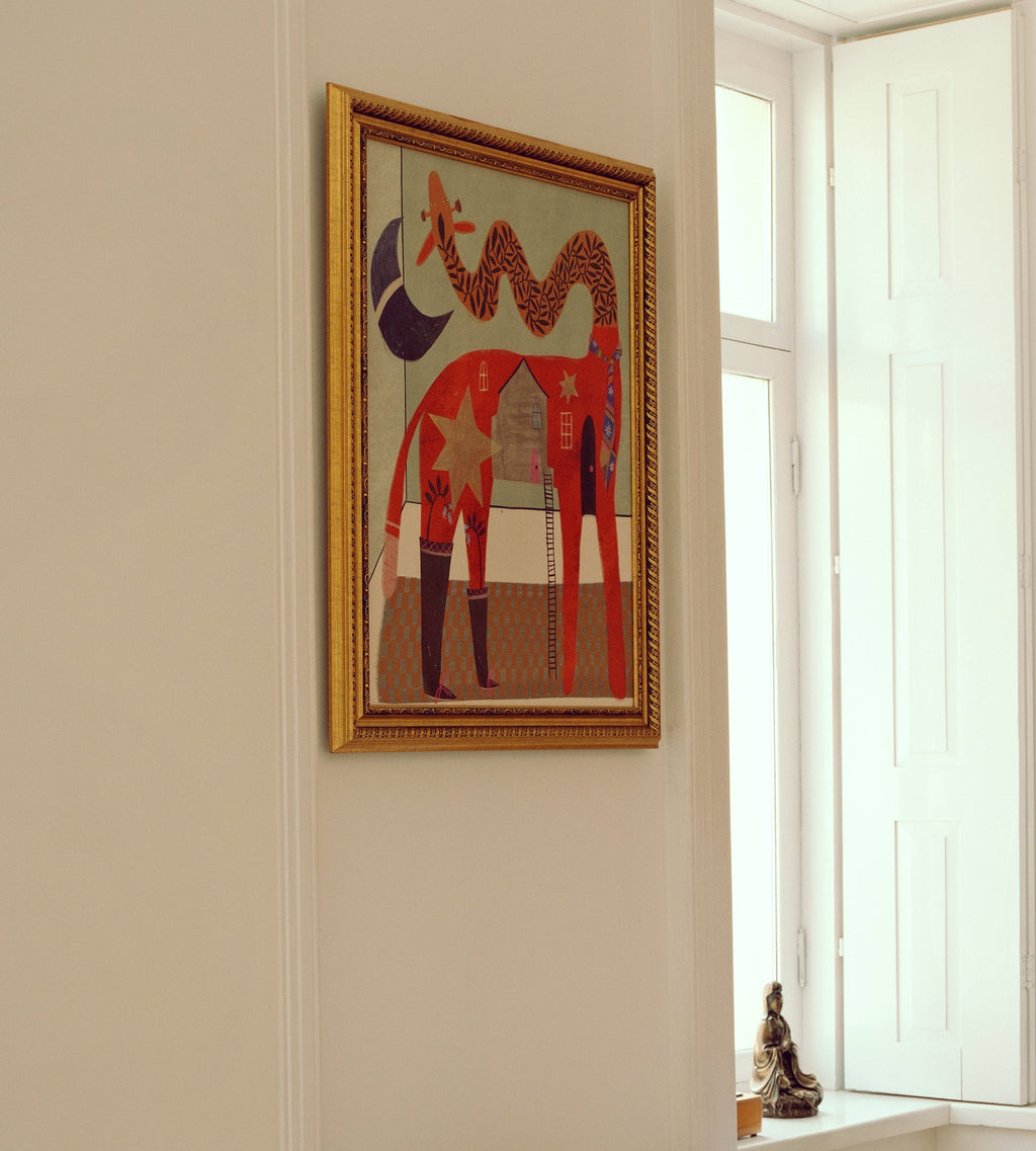 A framed art print featuring a stylized red giraffe with geometric patterns, hanging on a cream-colored wall next to a bright window.