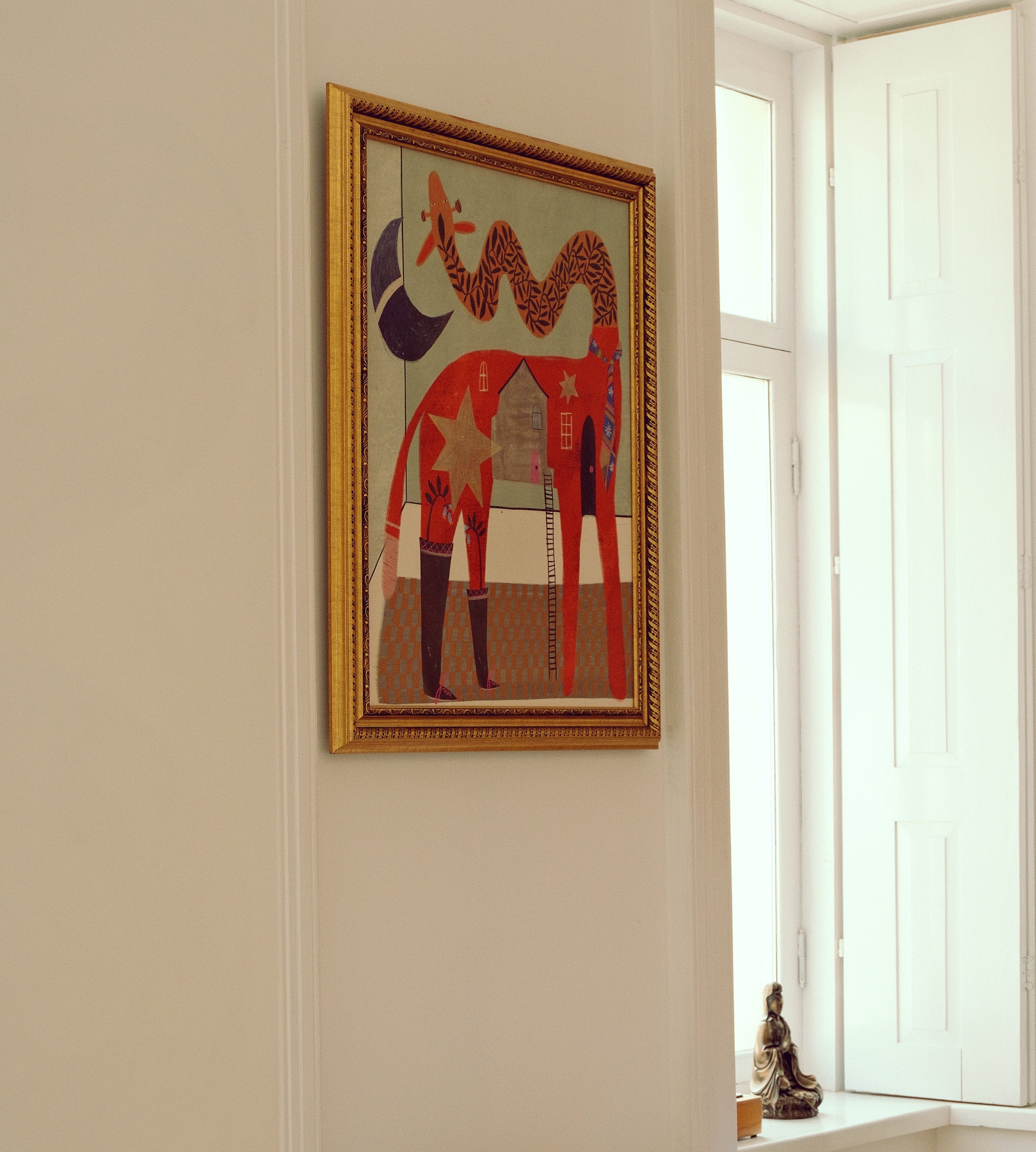 A framed art print featuring a stylized red giraffe with geometric patterns, hanging on a cream-colored wall next to a bright window.