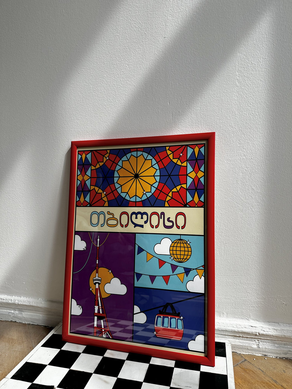 Illustrated “Tbilisi” poster by Mariam Kantaria in a wooden frame, colorful city-inspired artwork displayed on the floor in natural light.