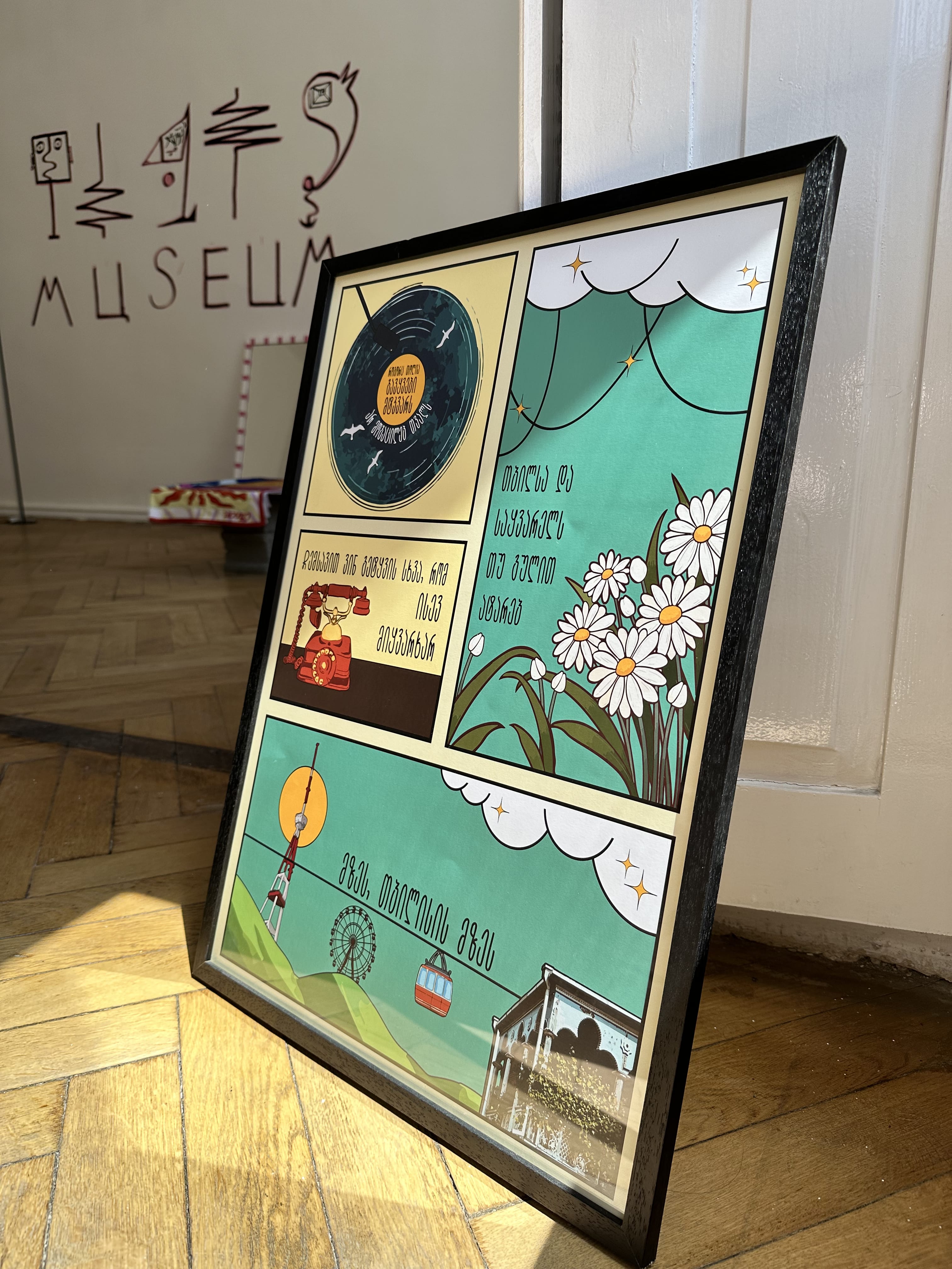 A framed poster featuring illustrated elements such as a record, a windmill, and flowers, with Georgian text, indicating a musical theme and cultural artwork.