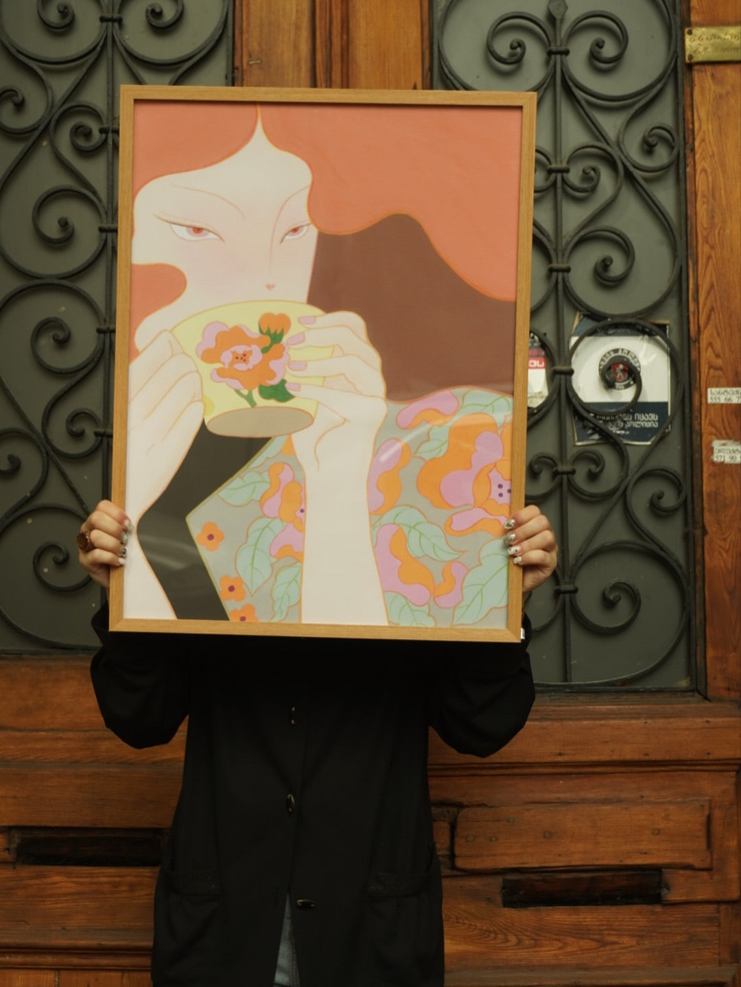 Person holding a framed artwork with a stylized illustration of a woman holding a vase against a decorative metal gate.