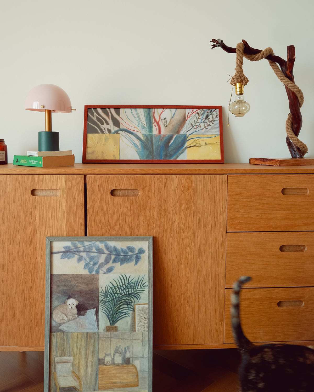 A styled interior scene showing a wooden sideboard with framed artwork leaning against it. A pink dome lamp sits on the left, a rope-wrapped light fixture stands on the right, and two framed abstract and botanical prints are displayed on and in front of the cabinet.
