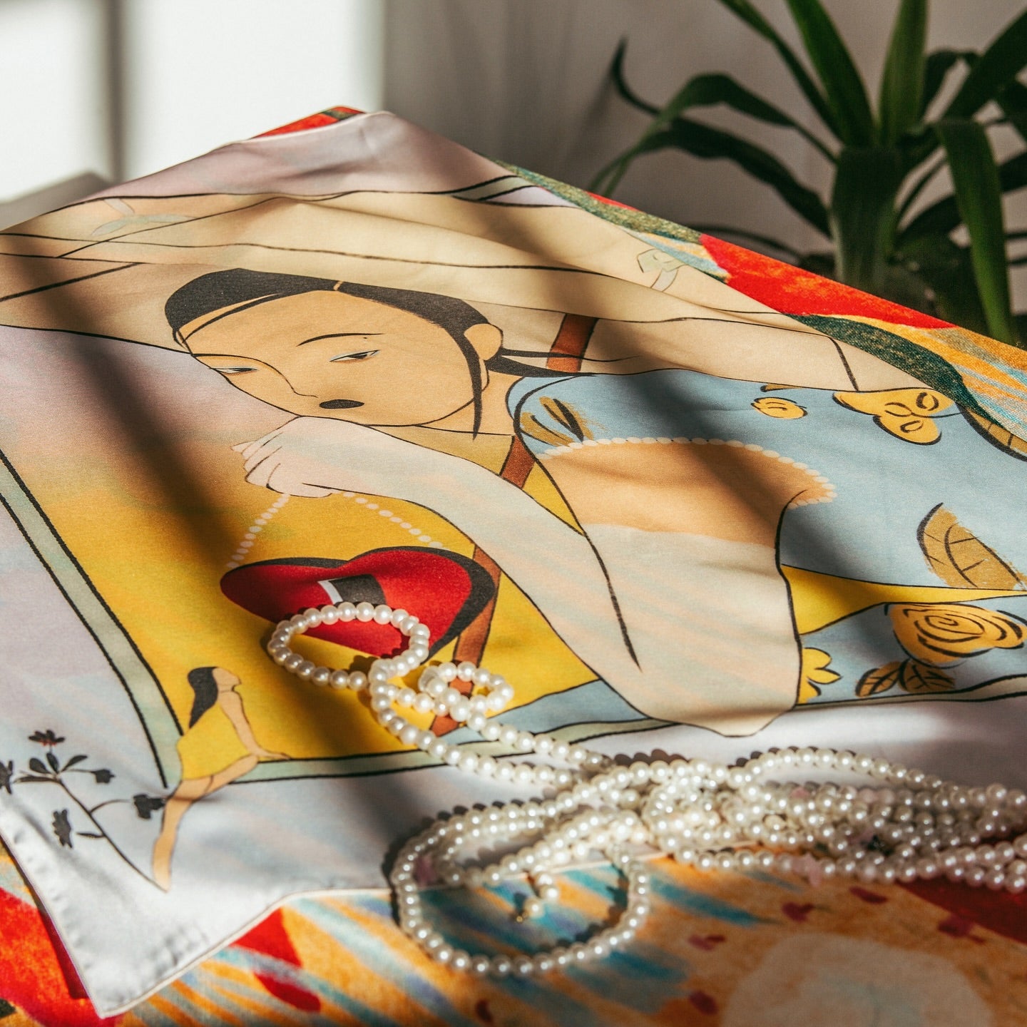 A silk scarf with an illustration featuring a woman and various objects, predominantly in pastel colors, with a strand of pearls lying on the scarf. Art silk scarf featuring poetic illustration by Lea Woo