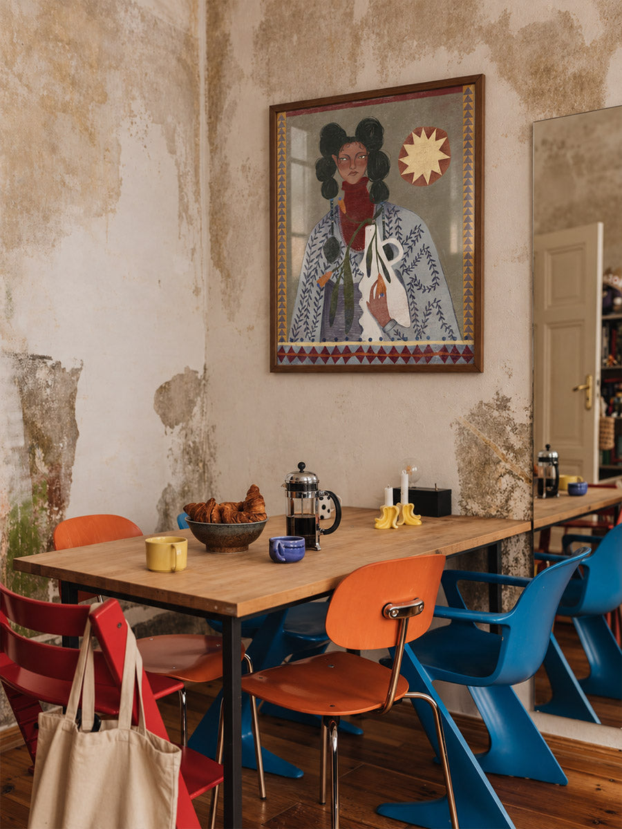 A framed portrait poster by DAE hangs on a distressed, textured beige wall above a wooden dining table with orange, blue, and red chairs in a rustic, industrial-style room.
