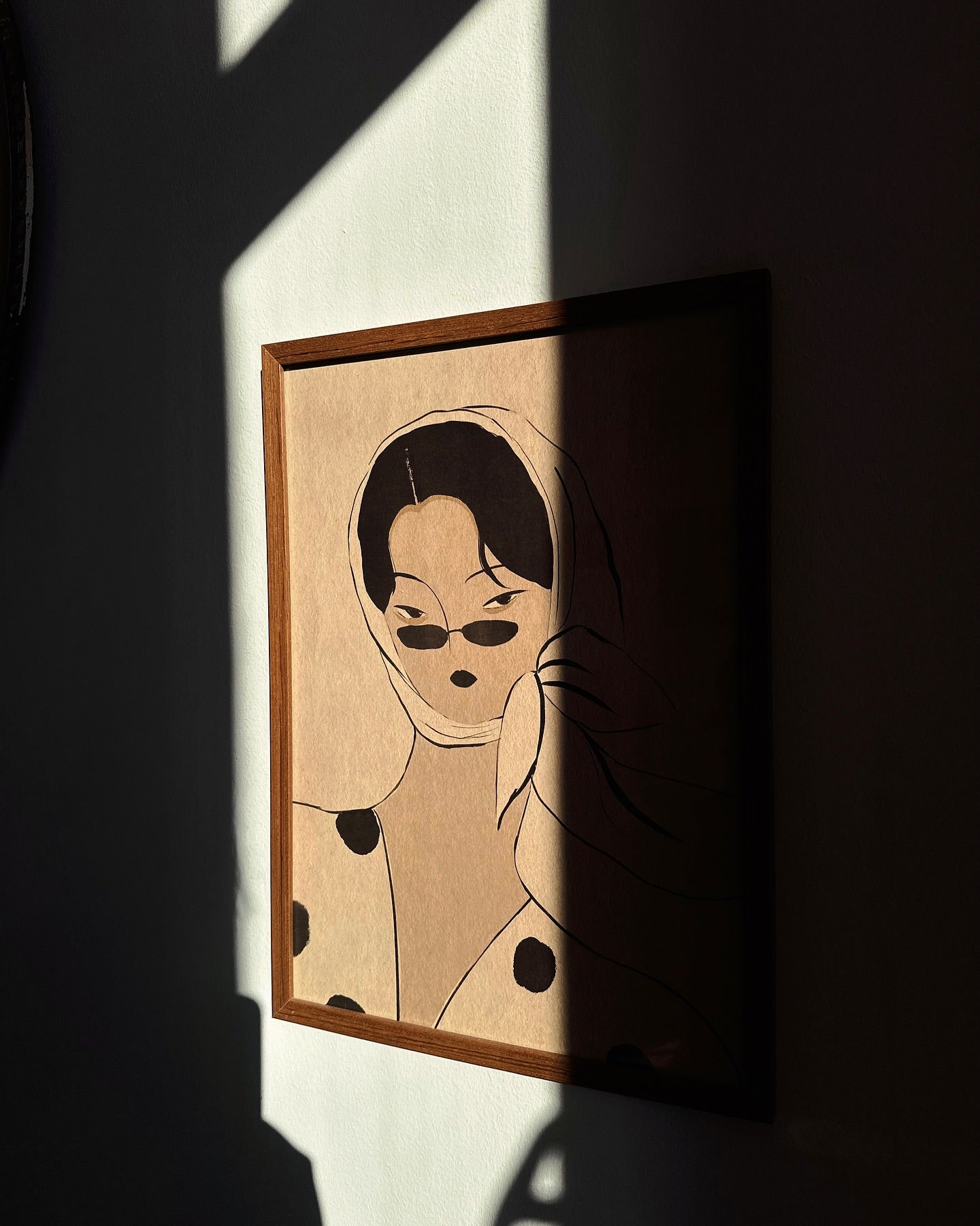A framed poster featuring a stylized illustration of a female figure with polka dots, displayed on a wall with natural lighting casting shadows.