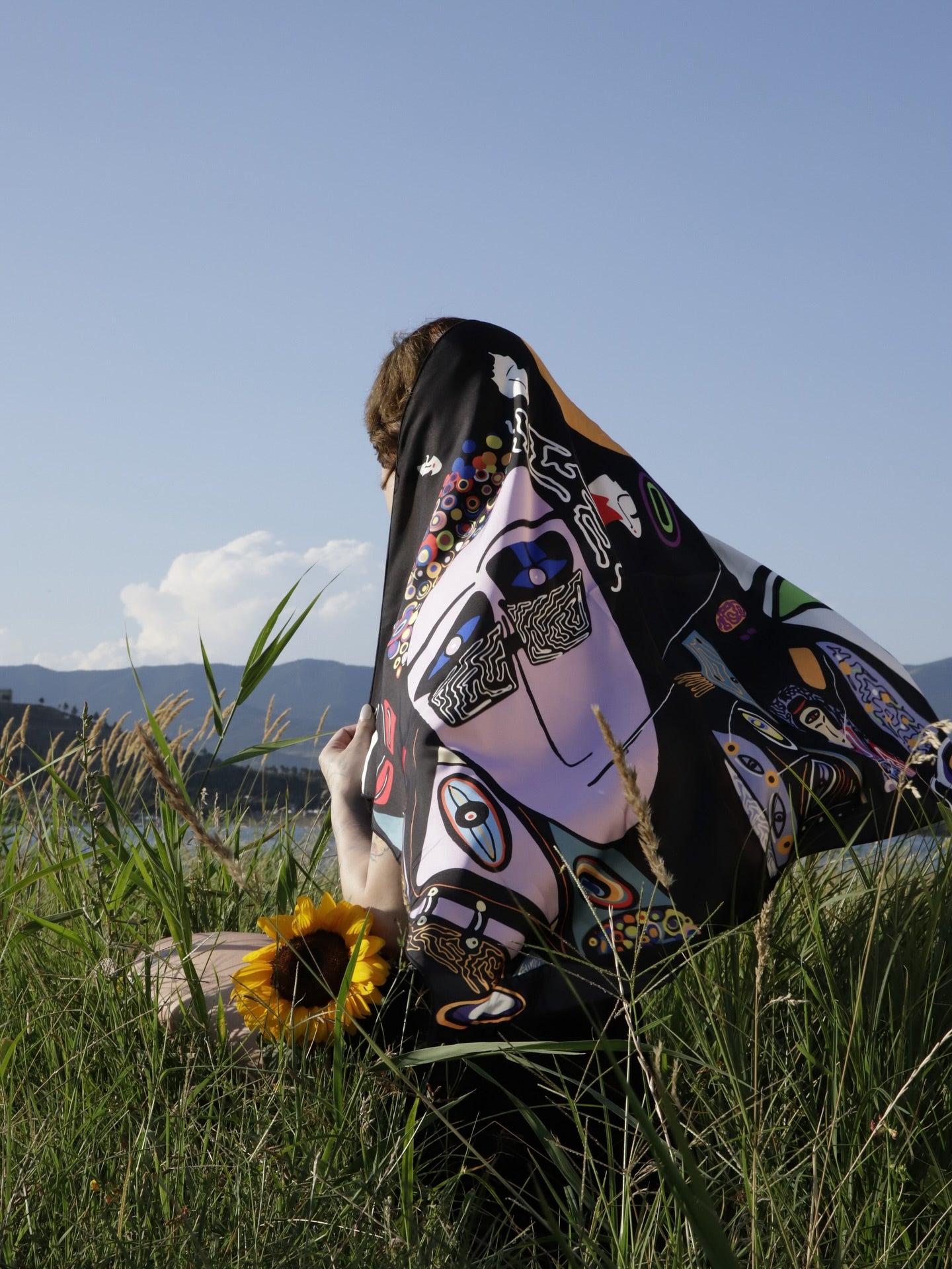 Person sitting in a field with a colorful scarf draped over their head. Scarf by DAE