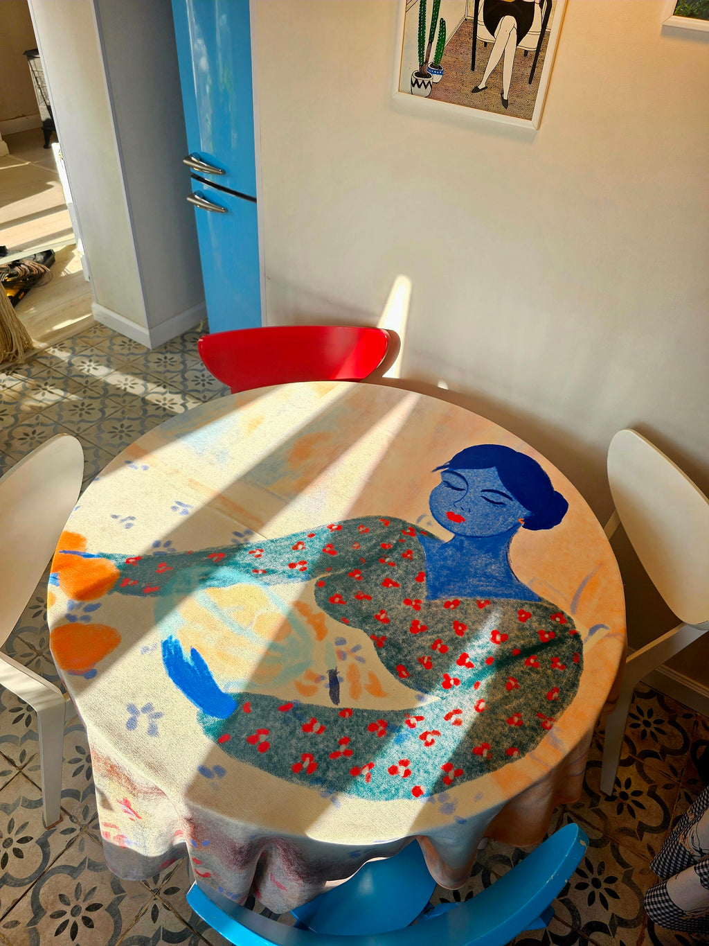 Round table with a colorful tablecloth featuring a woman's silhouette in a room with a blue cabinet and patterned floor.