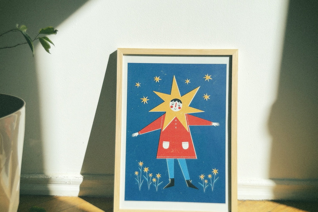 Framed illustration of a character with a star on a blue background, placed on a surface with a plant in the corner.